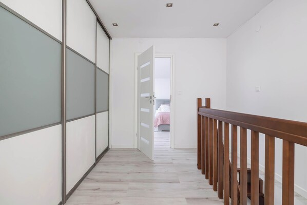 A hallway with a wooden railing, a door leading to a bedroom with a bed and a pink blanket, and a wall with a frosted glass panel.