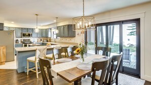 A modern kitchen with a central island, dining area with chairs, and a view of the outdoors through large sliding doors. - Swim, Fish & Boat: House on Stunning Higgins Lake! (Roscommon)