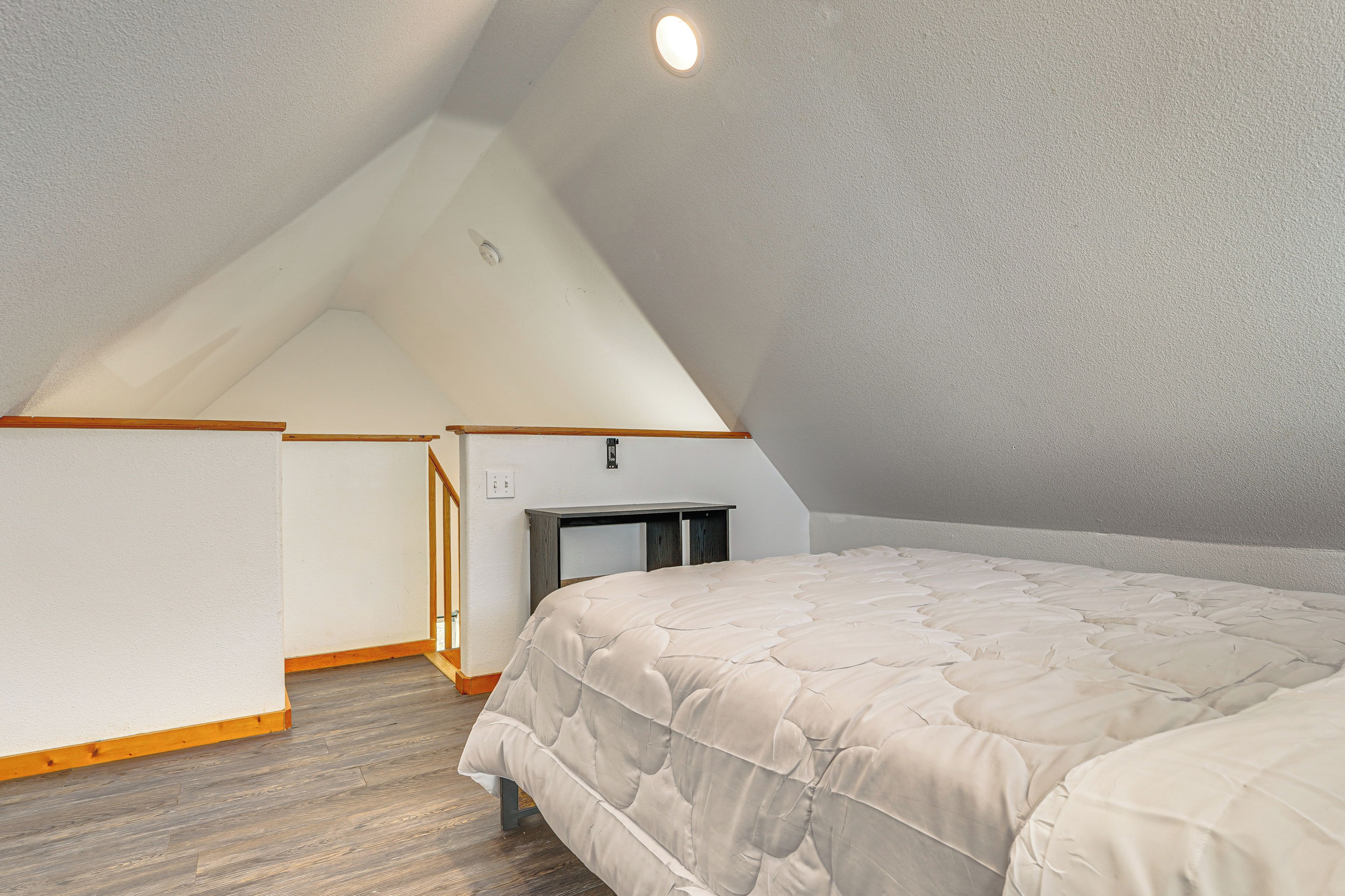 Quarto com teto inclinado, uma cama, uma mesinha e luminária fixada na parede.