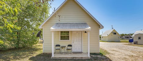 A small, single-story building with a gabled roof, a front door, and two chairs outside.