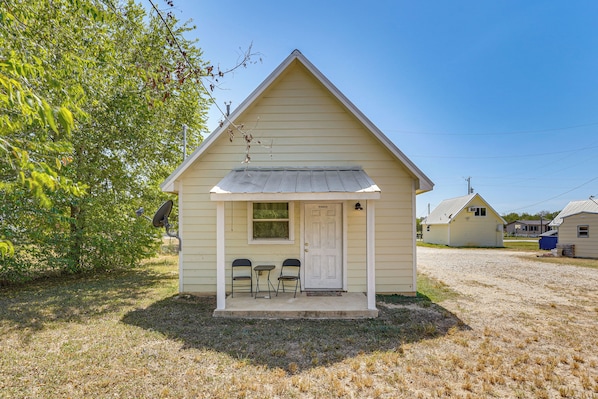 A small, single-story building with a gabled roof, a front door, and two chairs outside.