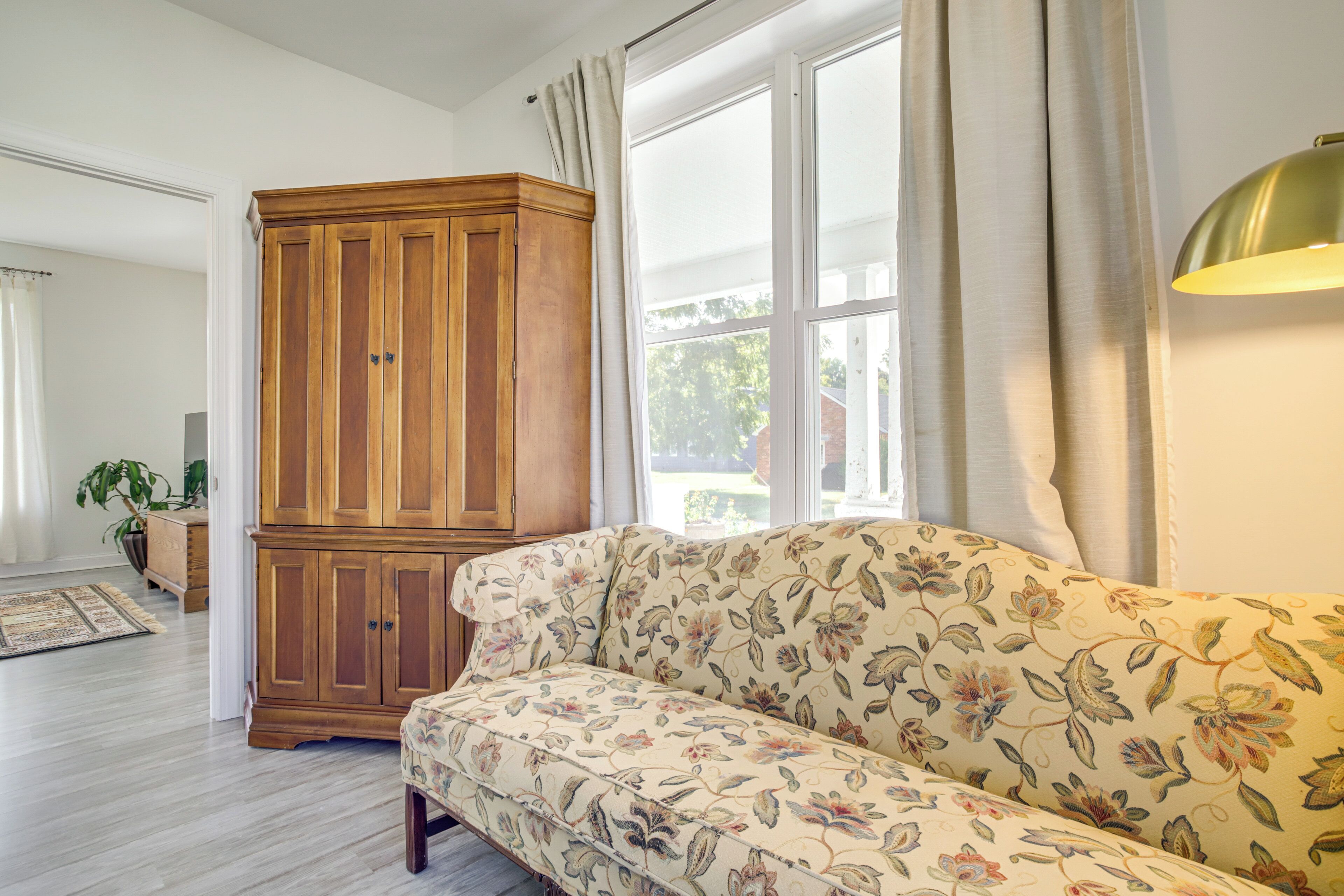 A living room with a floral sofa, wooden cabinet, and large windows with curtains.