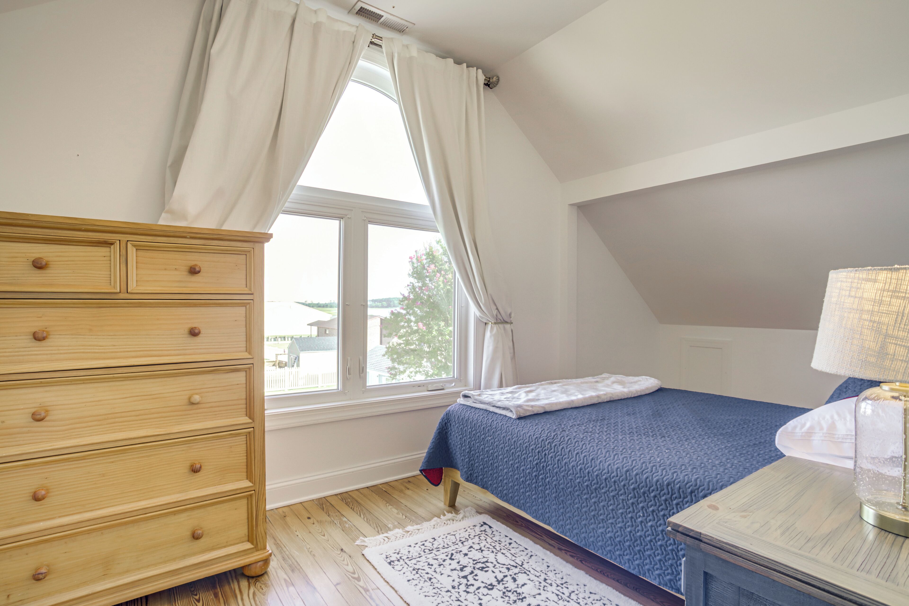 Une chambre à coucher avec une commode en bois, un lit recouvert d’une couverture bleue, une lampe blanche et une fenêtre avec des rideaux.