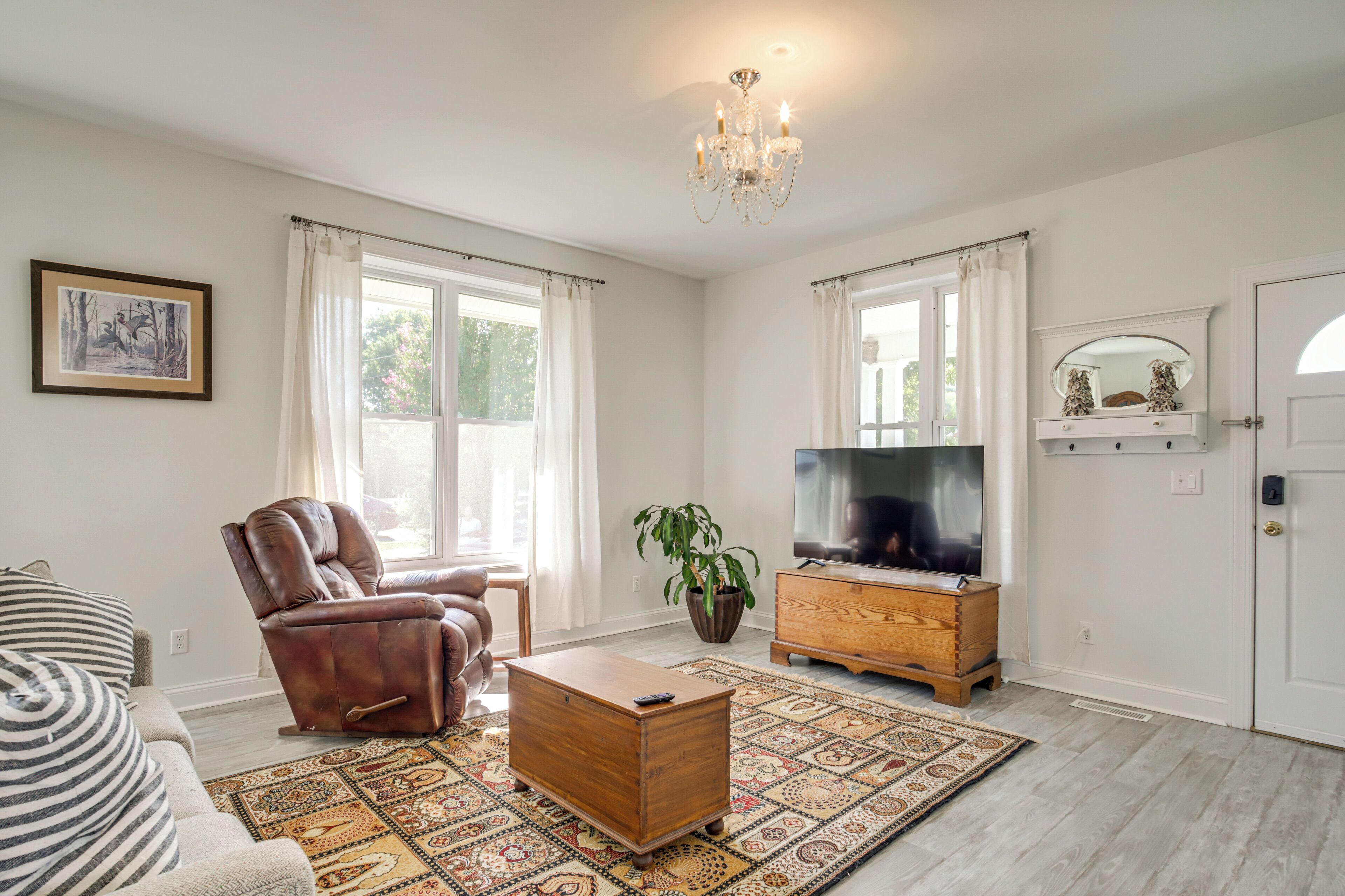 A living room with a leather armchair, a wooden coffee table, a television on a wooden stand, a patterned rug, and a chandelier.