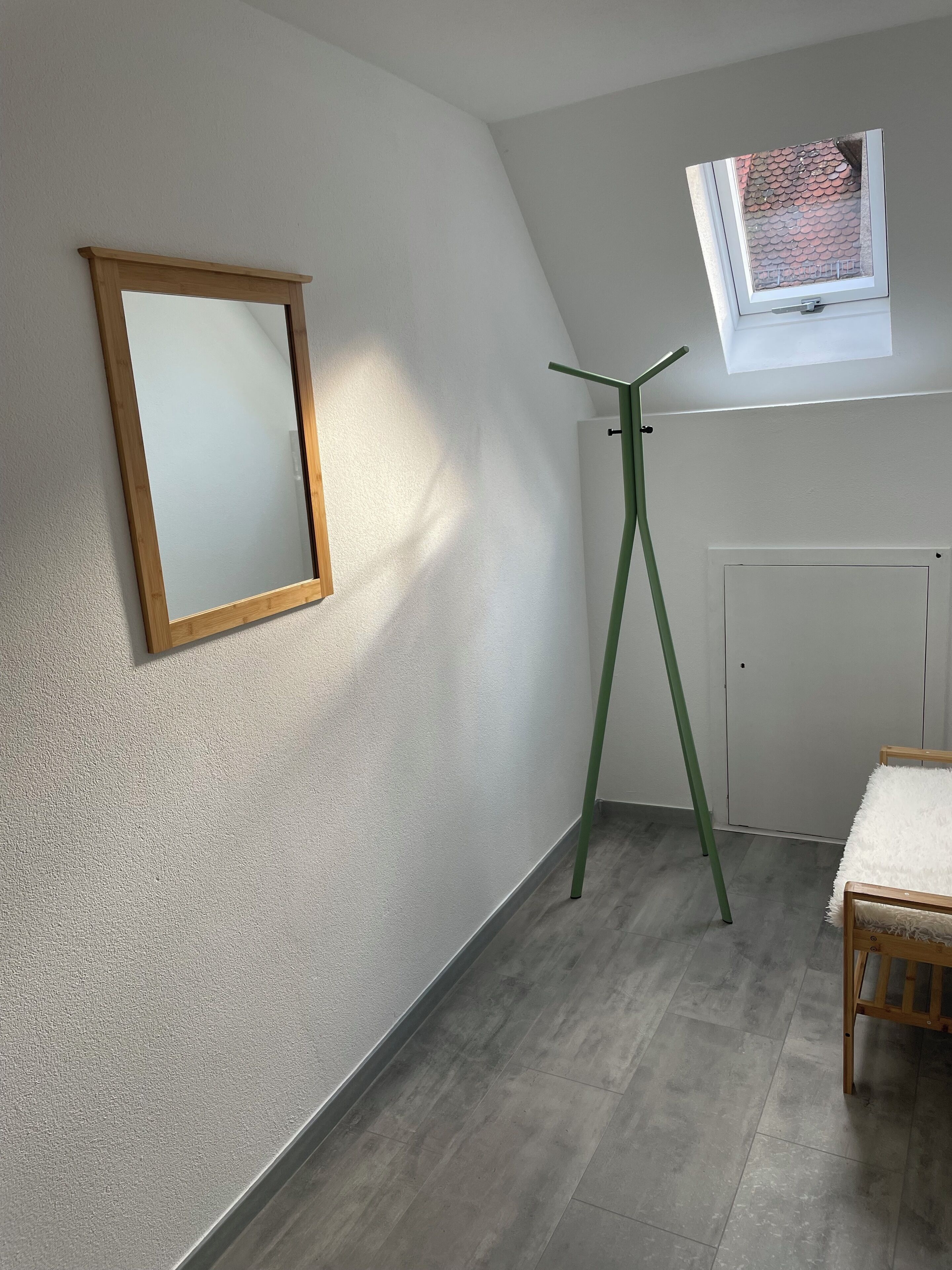 A room with a wooden-framed mirror, a green floor lamp, a white sofa, and a window with a red-tiled roof view.