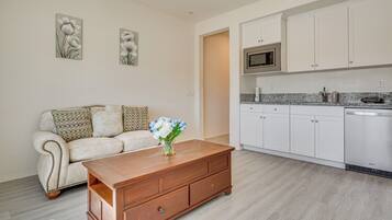 A modern living room with a wooden coffee table, a white sofa with patterned cushions, and a kitchen area with white cabinets and a built-in microwave.