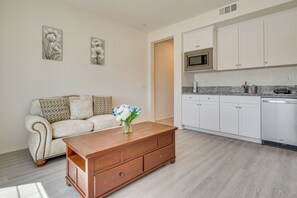 Un soggiorno moderno con un tavolino in legno, un divano bianco con cuscini decorati e un angolo cottura con mobili bianchi e forno a microonde integrato.