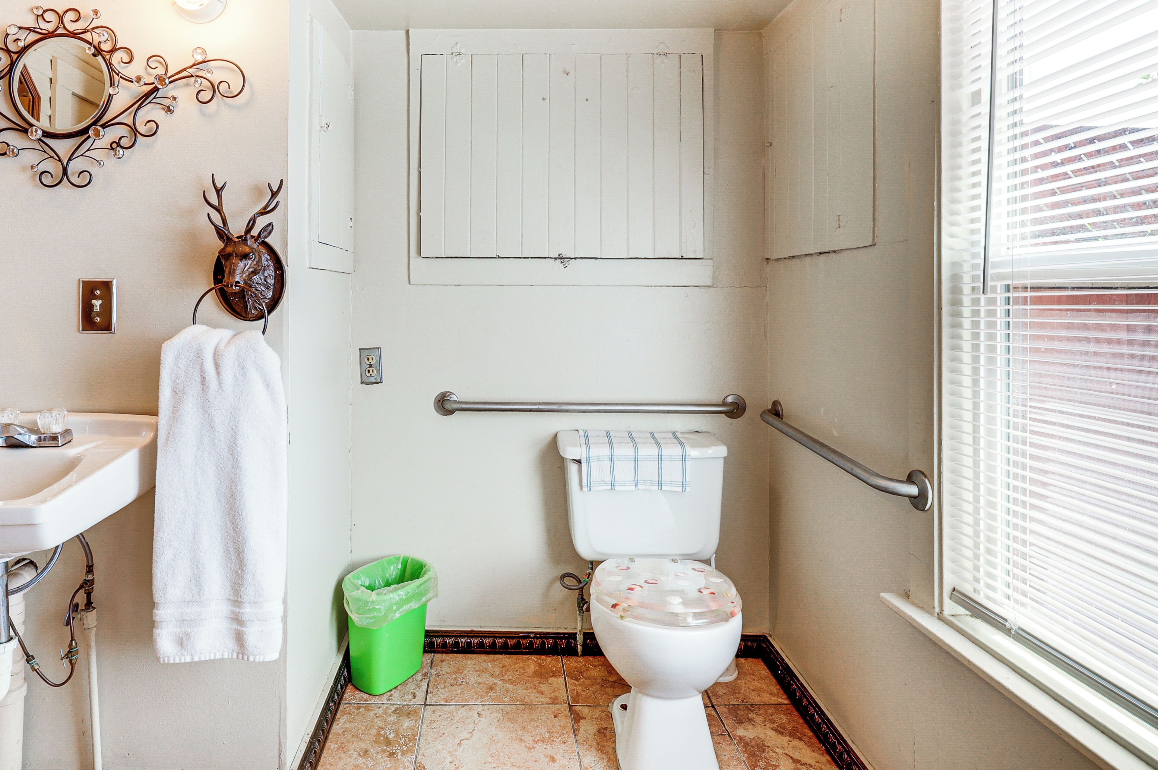 A bathroom with a white toilet, a sink, a green trash bin, a decorative deer head, and a window with blinds.