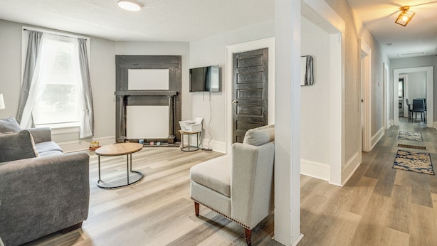 A modern living room with a sofa, armchair, and coffee table. There is a flat-screen TV mounted on the wall and a fireplace. The room has hardwood flooring and a large window with curtains.