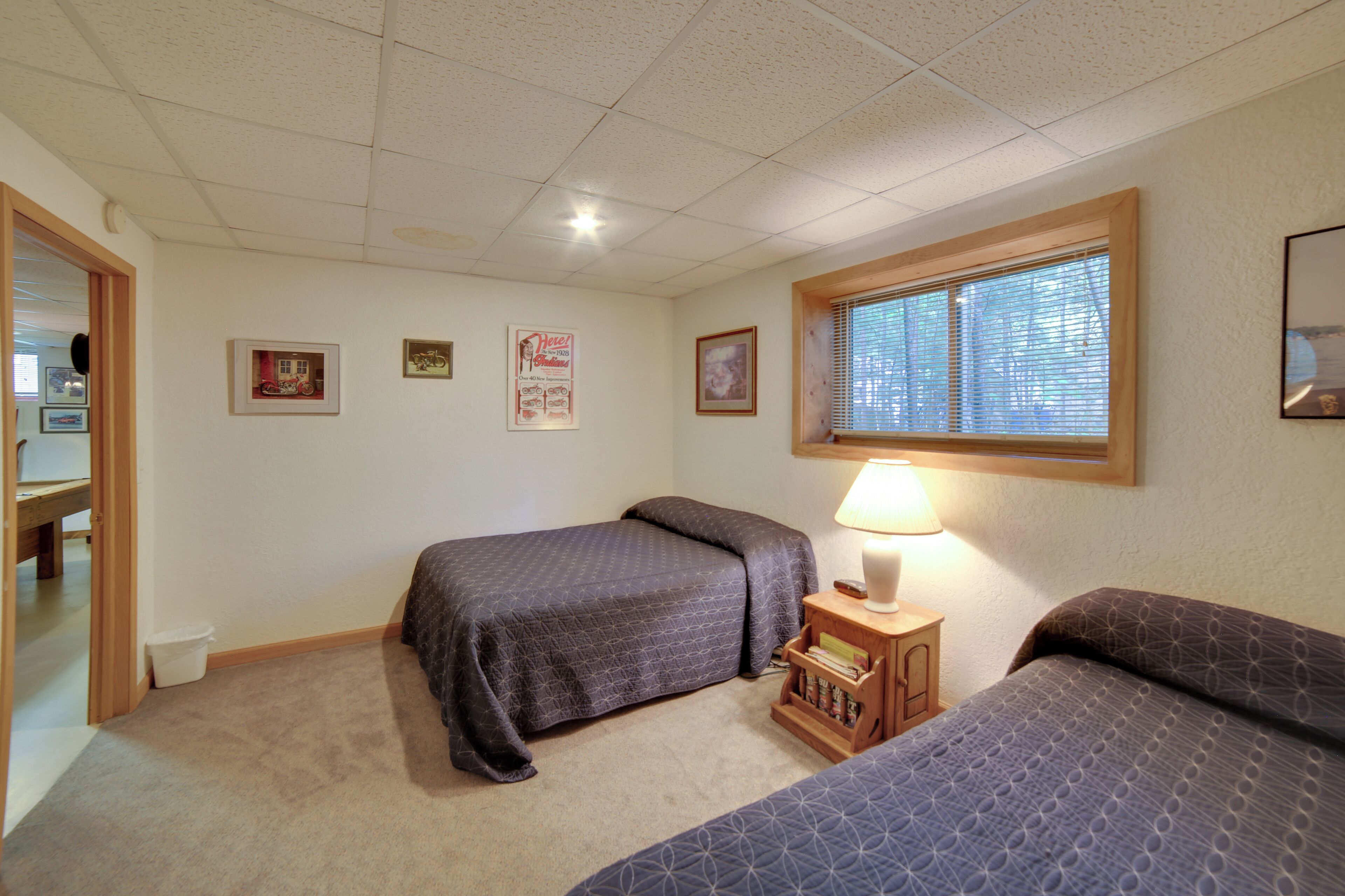 A bedroom with two beds, a window with blinds, a bedside table with a lamp, and a small shelf with books.