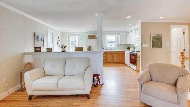 A spacious living room with a white sofa and a beige armchair, a kitchen in the background, and a large white column in the center.