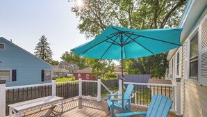 A wooden deck with a turquoise umbrella, two blue Adirondack chairs, and a white bench.