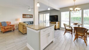 A spacious kitchen with a central island, dining area with wicker chairs, and a television in the background. - 3 Mi to Siesta Key Beach: Condo w/ Heated Pool! (Sarasota)