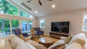 A spacious living room with a large TV, comfortable seating, and large windows offering a view of the outdoors.