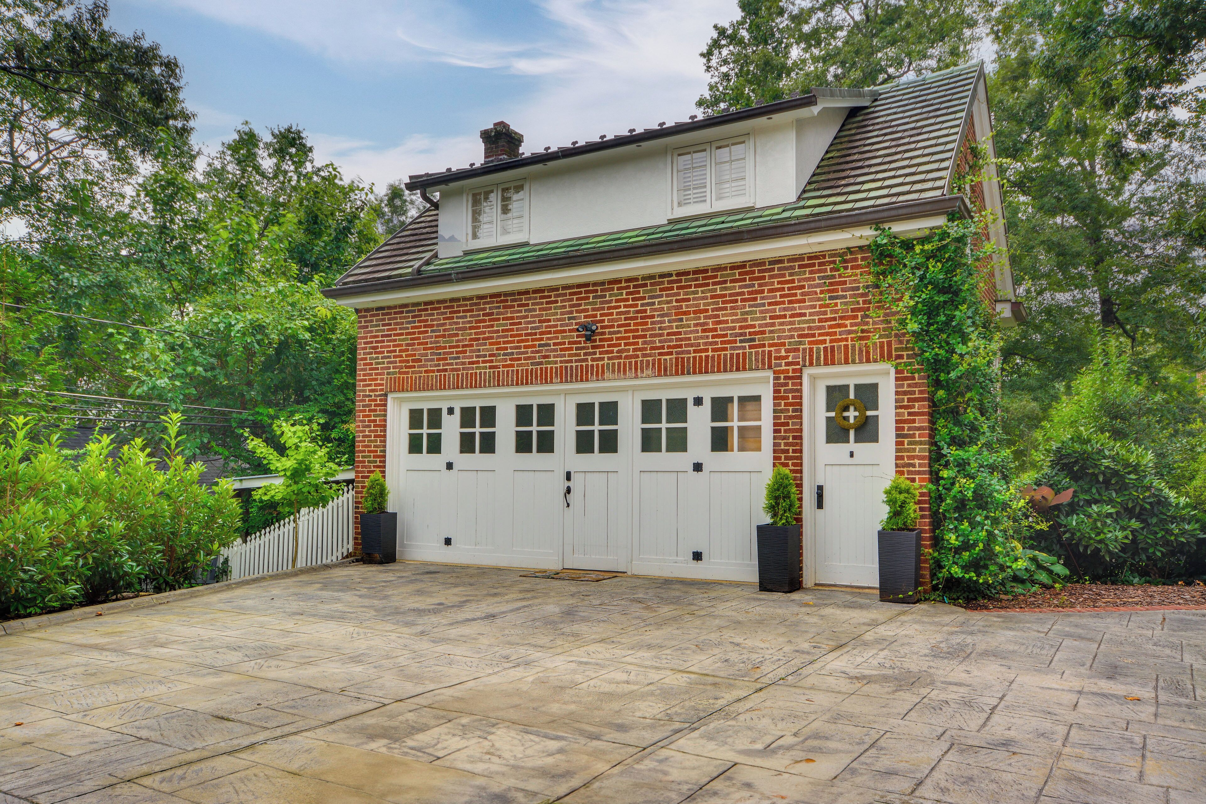 A two-story brick house with a garage, surrounded by lush greenery.