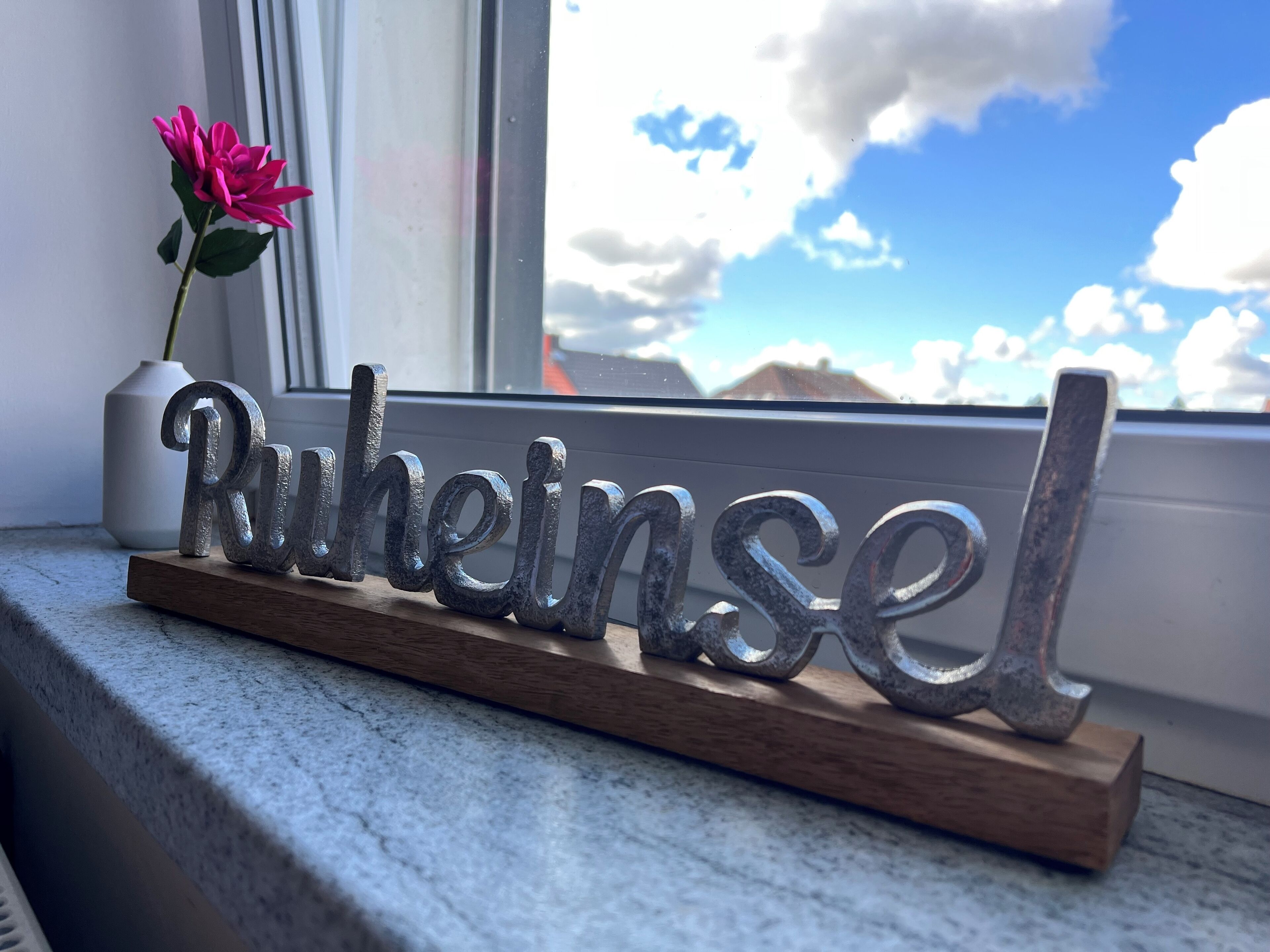 A metal sign spelling "Ruheinsel" on a wooden base, placed on a windowsill with a view of a cloudy sky and a pink flower in the background.