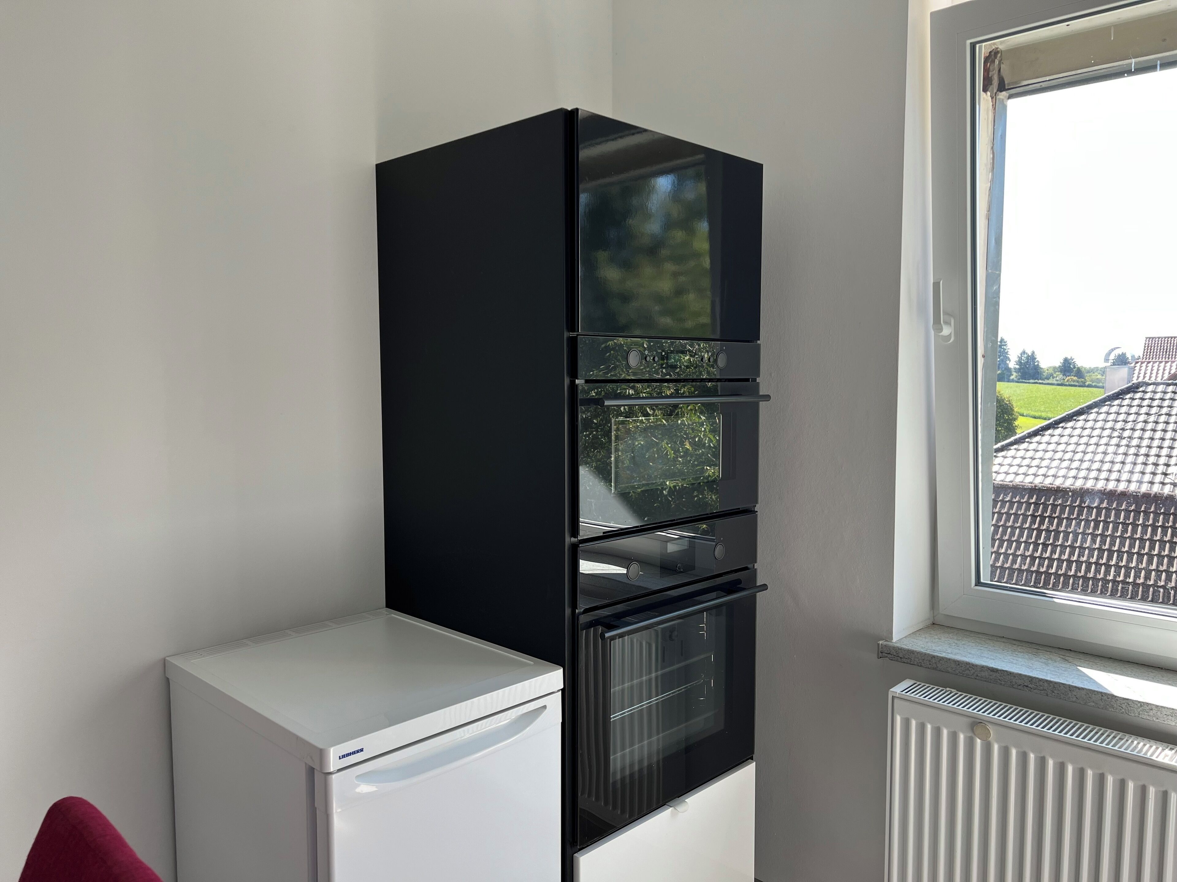 A compact kitchen with a white refrigerator, a black oven, and a window with a view of a tiled roof and greenery.