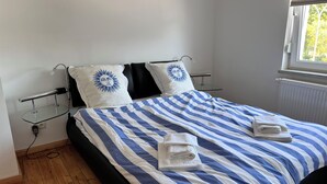 A neatly made bed with striped bedding and two pillows, a glass bedside table with a lamp, and a window with blinds.