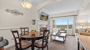 A dining area with a round table, chairs, and a bowl of fruit. There is a fireplace, a sofa, and a large window with a view.