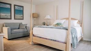 Ein Bett mit Himmelbett, ausgestattet mit weißer Bettdecke und Kissen, ein graues Sofa und zwei gerahmte Strandbilder an der Wand.