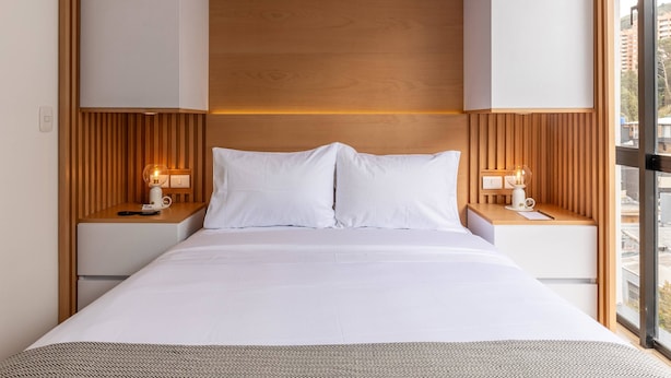 Een hotelkamer met een groot bed, een houten hoofdbord, twee nachtkastjes met lampen, en uitzicht op gebouwen door een groot raam.