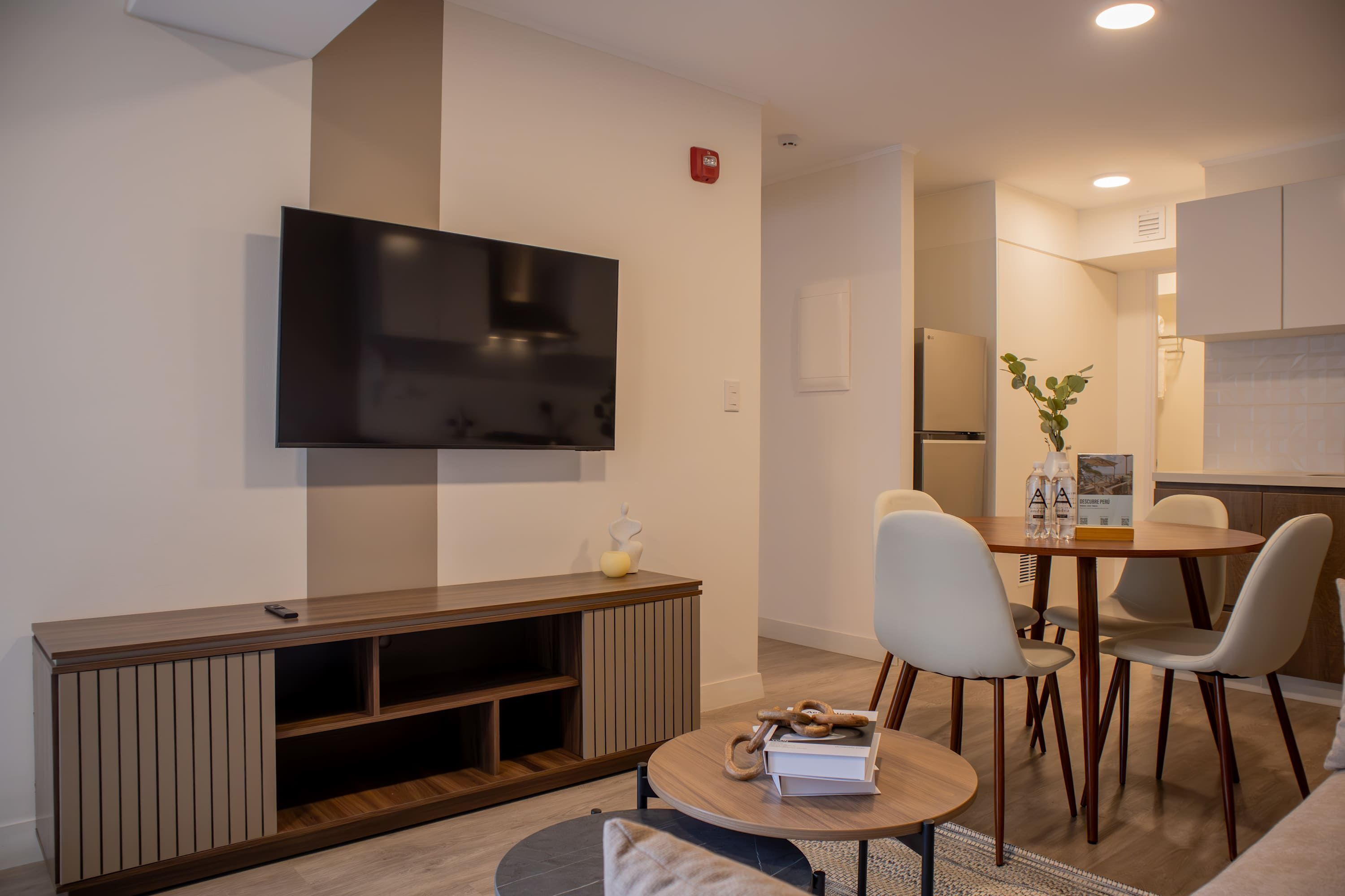 A modern living room with a flat-screen TV, a wooden entertainment unit, a round dining table with chairs, and a kitchen area in the background.