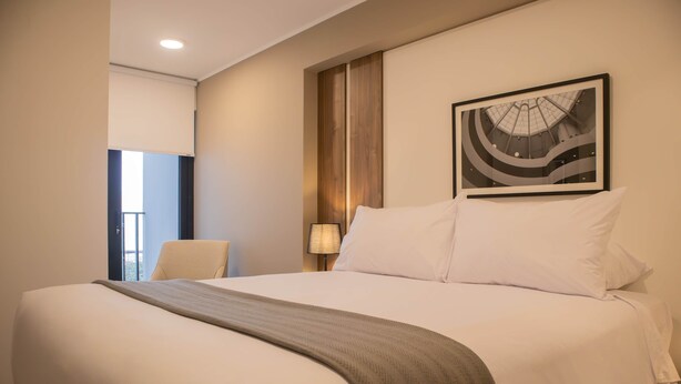 Quarto de hotel com uma cama grande, uma poltrona, um abajur e um quadro na parede.