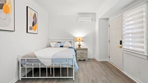 A bedroom with a white metal bed frame, a bedside table with a lamp, a wall-mounted air conditioner, and a door with blinds. - Near LA Cozy Home With Full Kitchen Laundry (San Gabriel)