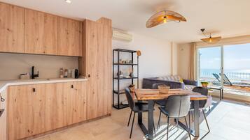 Una cocina moderna con armarios de madera, un comedor con mesa y sillas de madera, una estantería negra y vistas al mar a través de amplios ventanales.