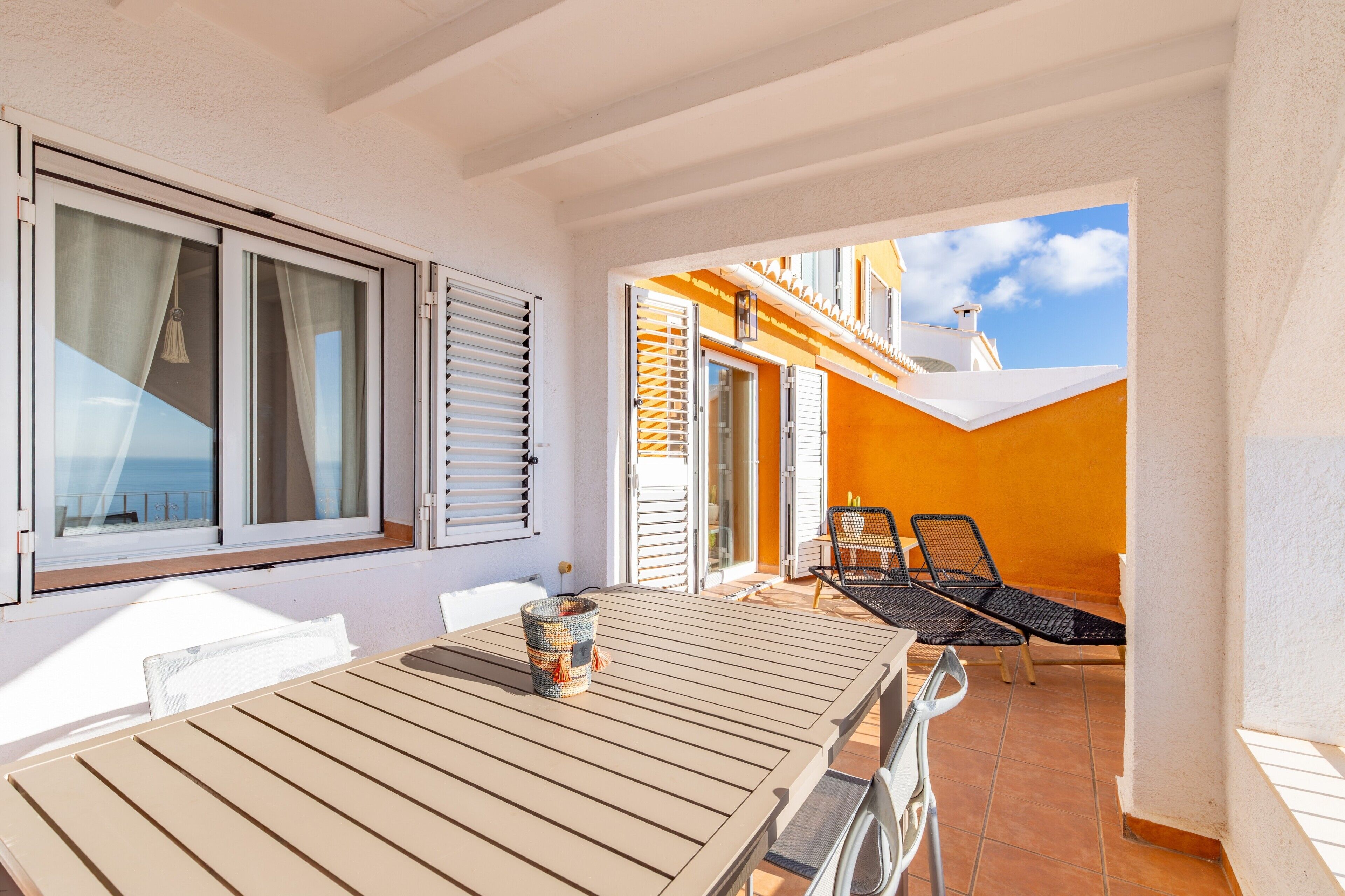 A sunlit patio with a table and chairs, a view of the sea through a window, and a building with orange walls.