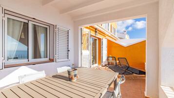 A sunlit patio with a table and chairs, a view of the sea through a window, and a building with orange walls.