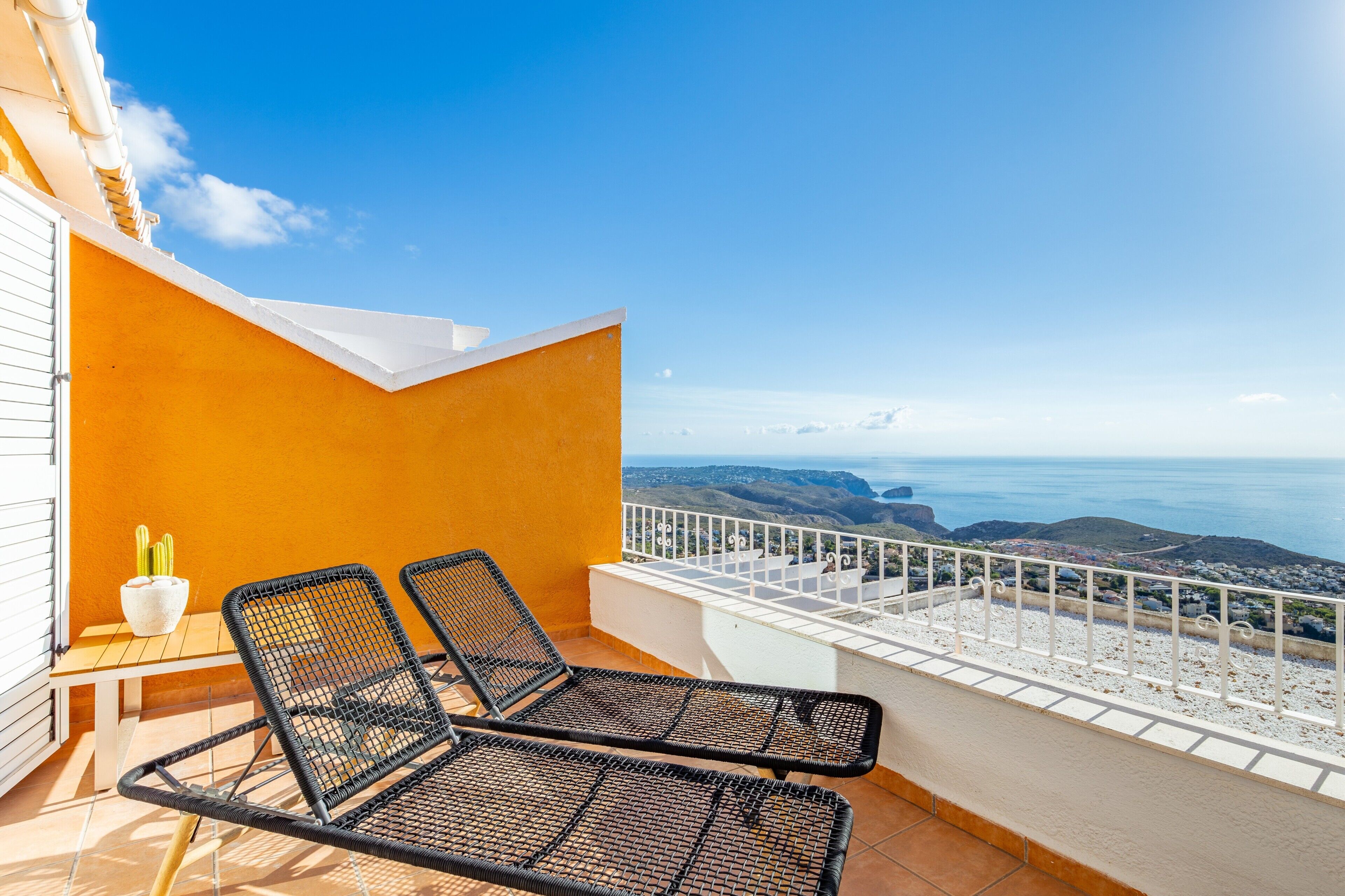 Ein Balkon mit zwei schwarzen Korbstühlen, einem kleinen Tisch mit einem Blumentopf und einem weißen Geländer mit Blick auf eine Küstenlandschaft.