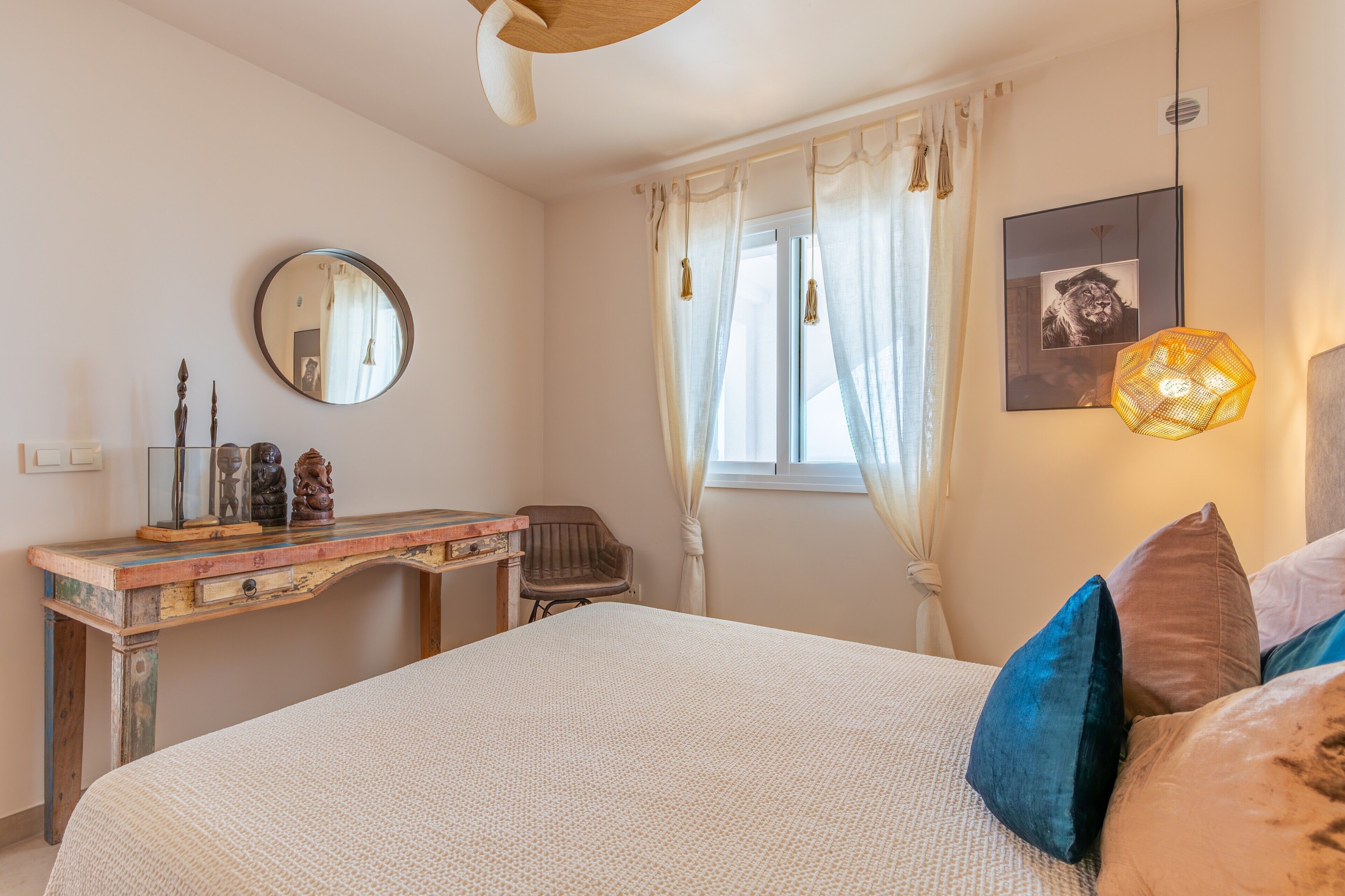A bedroom with a bed, a desk, a chair, a mirror, a window with curtains, and a wall-mounted light fixture.