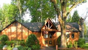 Une cabane en rondins dotée d’une cheminée centrale, entourée d’une pelouse bien entretenue et d’une allée menant à l’entrée.