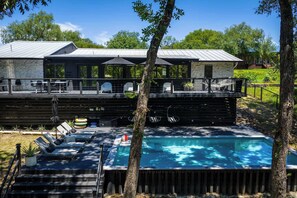 Un área de piscina moderna al aire libre con sillones de descanso y sombrillas, rodeada por una terraza de madera y muros de piedra.