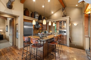 A kitchen with a central island, bar stools, hanging pendant lights, and wooden beams.