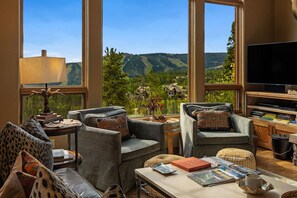 Un salón acogedor con un amplio ventanal que ofrece una vista panorámica de colinas y árboles. Hay un televisor de pantalla plana, una mesa de centro con libros y revistas, y un mobiliario cómodo.