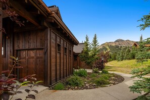 A wooden cabin with a stone pathway leading to the entrance, surrounded by a well-maintained garden and a mountain view.