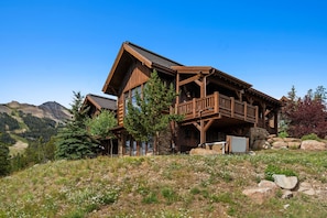 A wooden cabin with a balcony, surrounded by mountains and greenery.
