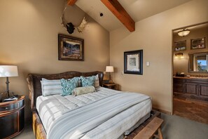 A bedroom with a large bed, wooden headboard, two bedside tables with lamps, a framed picture on the wall, and a wooden bench at the foot of the bed.