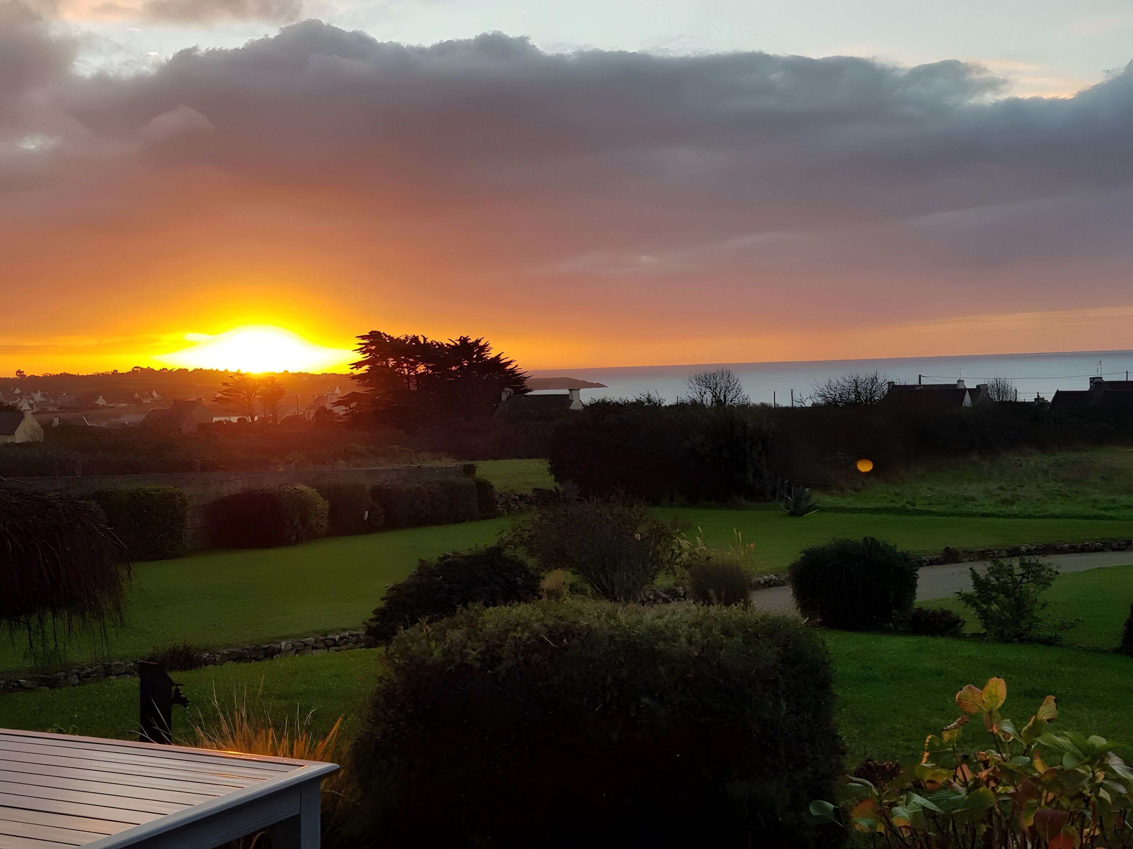 A sunset over a residential area with a view of the sea, a grassy lawn, and a stone wall.