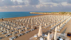 Une plage bordée de rangées de chaises longues et de parasols, une terrasse en bois avec des tables et des chaises, et une côte rocheuse en arrière-plan.