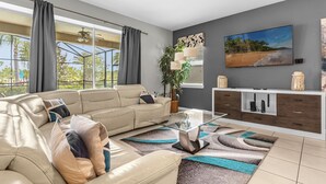Ein modernes Wohnzimmer mit einem beige farbenen Sofa, einem Glastisch und Blick auf Palmen durch große Fenster.