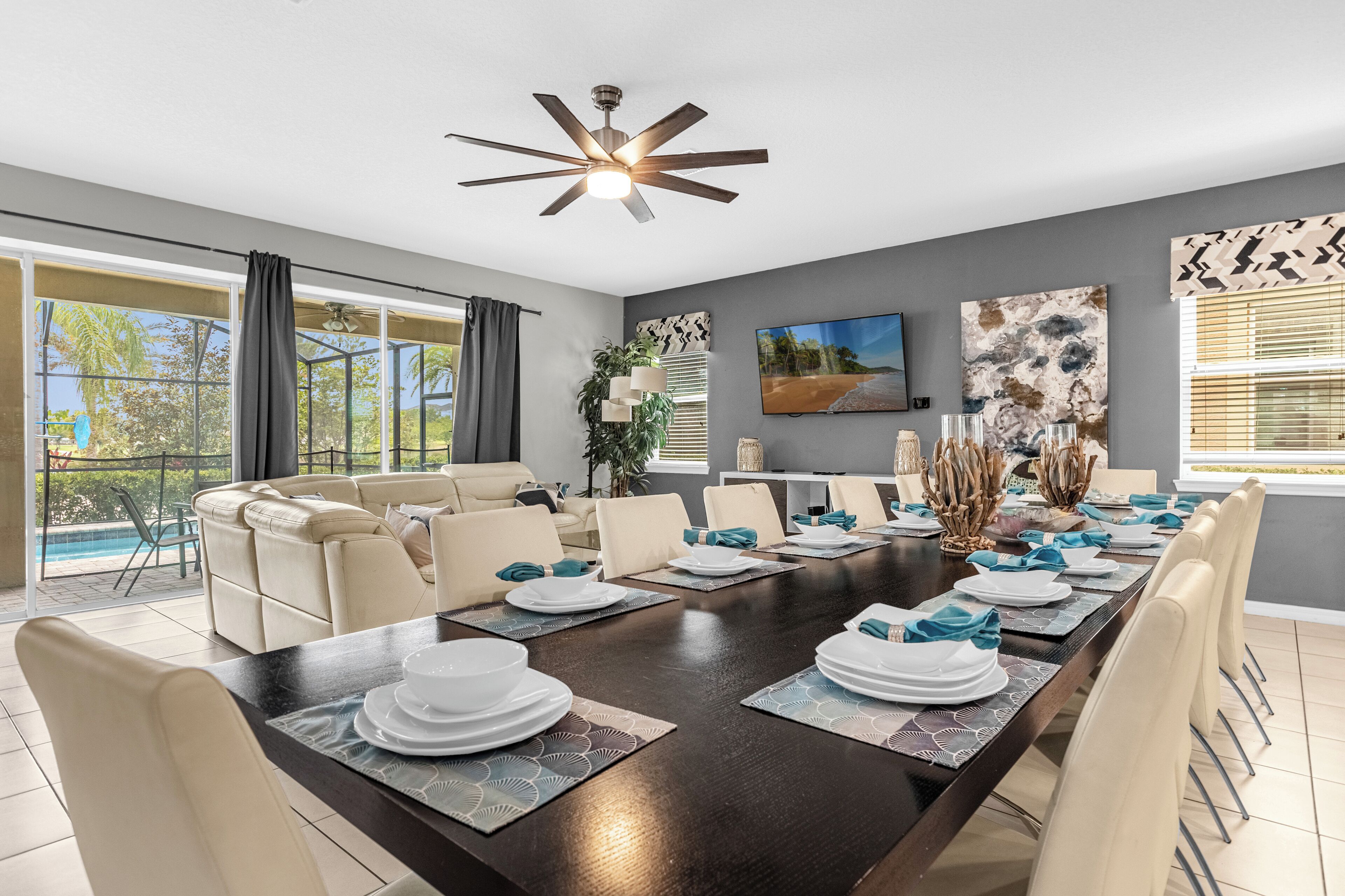 Un comedor con una larga mesa dispuesta para seis personas, rodeada de sillas color beige, con un ventilador de techo arriba y un gran ventanal a un lado.