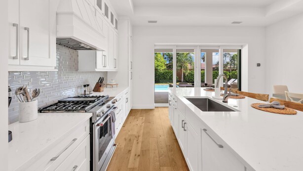 A modern kitchen with white cabinetry, a large island, and a view of a pool through glass doors. - Beach Days, Sunset Skies & Poolside Living (Naples)