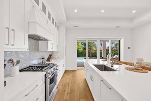 A modern kitchen with white cabinetry, a large island, and a view of a pool through glass doors. - Beach Days, Sunset Skies & Poolside Living (Naples)