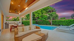 A modern outdoor living area with a pool, wicker furniture, and a view of lush greenery. - Beach Days, Sunset Skies & Poolside Living (Naples)