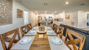 A dining area with a wooden table set for six, surrounded by wooden chairs. A kitchen is visible in the background.