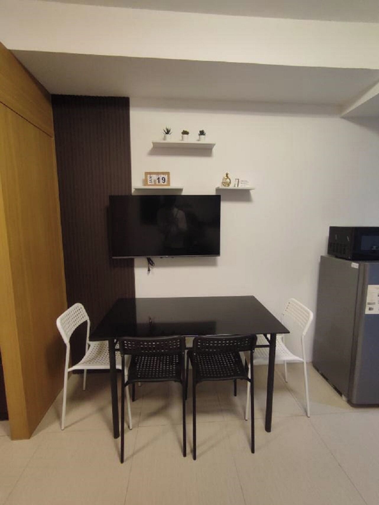 A small dining area with a black table and four chairs, a TV mounted on the wall, and a refrigerator to the right.