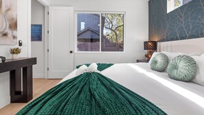 A bedroom with a large bed, a nightstand, a desk, and a window with a view of a house.